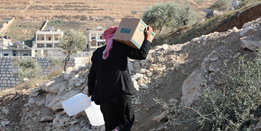 In Syria, a man carries a box on his right shoulder and two jugs of water in his left hand.