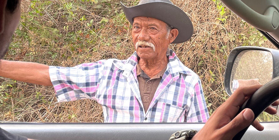 A farmer stands outside of a vehicle pointing at something in the distance. 
