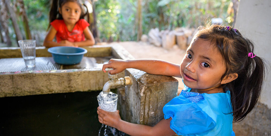 In Honduras, a little girl pours fresh clean water from a tap into a cup while another little girl watches in the distance.